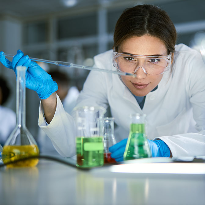 Eine Frau beugt sich über einen Tisch voller Bechergläser mit farbigen Flüssigkeiten und gibt mit einer Pipette ein paar Tropfen in einen Erlenmeyerkolben.